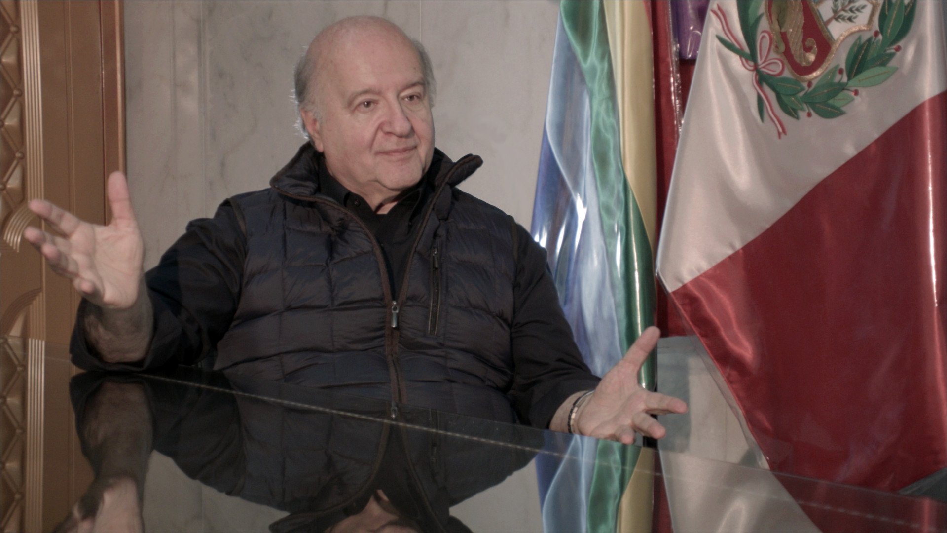 Disparity Film Still, Hernando de Soto sitting at table with Peruvian flag behind him